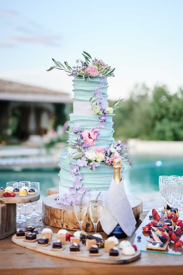 Close-up detail shot of an outdoor wedding dessert table set beside a pool, with a property building visible in the background. The centerpiece is a tall four-tier wedding cake with a mint green ruffled fondant finish, decorated with a cascading arrangement of lavender, pink lisianthus, white ranunculus, and olive branch greenery, placed on a raw wood slice stand. Two champagne flutes and a bottle of champagne are positioned in front of the cake on the wooden table. The surrounding spread includes individual petit fours and miniature desserts arranged on wooden boards, a tiered stand with small fruit-topped pastries, a berry and fresh fruit tart platter, and rows of crystal glassware. The overall dessert table styling follows a rustic-meets-garden-party aesthetic with a pastel color palette of mint, lavender, and pink.