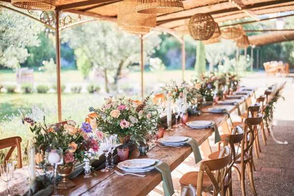 A wide shot of a long wooden farmhouse dining table set for a wedding reception under an open-sided pergola or covered outdoor terrace, with a garden visible in the background. The table is dressed with a lush floral runner featuring mixed blooms in pink, orange, purple, and yellow tones with abundant greenery, arranged in copper and terracotta vessels alongside white taper candles. Place settings include light blue-grey charger plates, crystal glassware, and cloth napkins, with cross-back wooden chairs and sage green upholstered chairs alternating along the sides. Woven rattan pendant lights hang from the pergola ceiling, and the overall styling aesthetic is bohemian and rustic with warm, earthy tones.