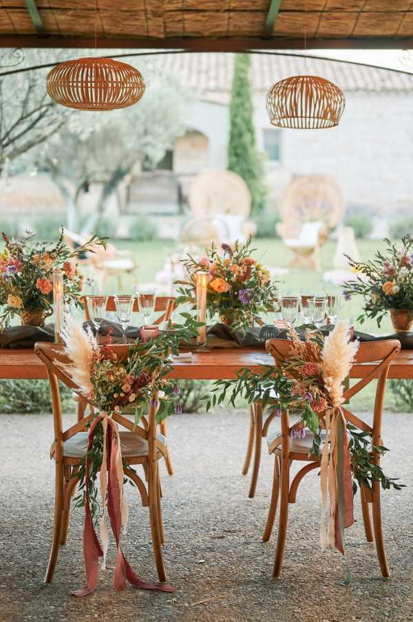 A detail shot of a styled wedding reception tablescape set under an open-sided covered terrace, with a Provençal-style stone building and garden visible in the background. The wooden farm table is dressed with charcoal grey linen runners, clear glassware, and tall amber candleholders, flanked by floral centerpieces in terracotta pots featuring burnt orange, rust, peach, and purple blooms with lush greenery. The two sweetheart chairs at the near end are decorated with lush garlands of olive-toned foliage, pampas grass, and small rust and peach flowers, tied with trailing dusty rose and terracotta silk ribbon. Two rattan dome pendant lights hang overhead from a thatched-style ceiling, reinforcing a warm, boho-rustic aesthetic with an autumnal color palette throughout.