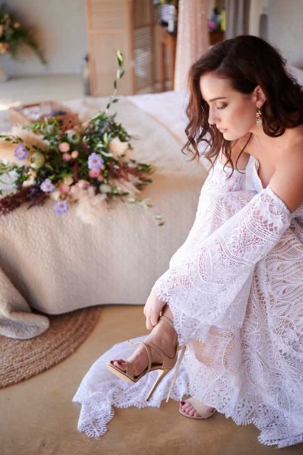 A bride is photographed indoors during the getting-ready phase, crouching down to fasten a nude strappy stiletto heel. She wears a white off-the-shoulder lace wedding dress with wide bell sleeves and a scalloped hem, featuring an intricate geometric lace pattern. Her dark wavy hair falls loosely around her shoulders and she wears gold drop earrings. In the background, a bridal bouquet rests on a cream textured surface over a jute rug, featuring a loose arrangement of lavender, blush, peach, and ivory blooms mixed with dried pampas grass and greenery, suggesting a boho styling theme. The shot is a close-up portrait taken from a slightly elevated angle.