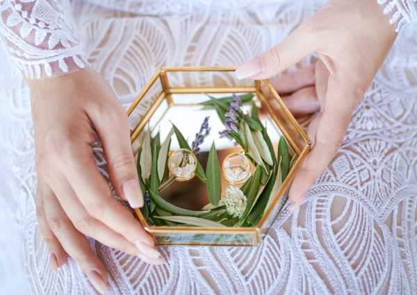 A close-up detail shot of a bride's hands holding a hexagonal gold and glass geometric ring box containing two wedding bands resting on a bed of greenery, including eucalyptus leaves and small sprigs of lavender. The rings appear to be gold bands, and the mirrored base of the box reflects the arrangement. The bride is wearing a white lace long-sleeve dress with an intricate patterned lace overlay, and her nails are finished with a neutral manicure. The styling has a modern boho aesthetic with the geometric brass box and botanical ring display.