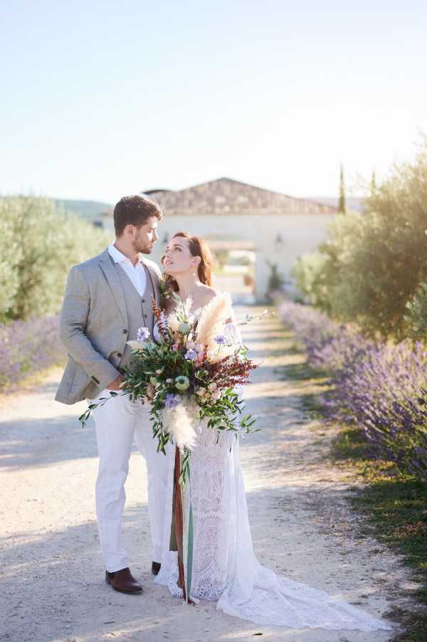 A couple portrait taken outdoors on a gravel path lined with lavender at golden hour, with a stone Provençal farmhouse visible in the background. The bride wears an off-shoulder white lace boho-style gown with a train and holds a large, loose bouquet featuring pampas grass, burgundy foliage, purple and blue wildflowers, peach blooms, and trailing green ribbons. The groom is dressed in a light grey linen blazer with a waistcoat, white trousers, and brown leather shoes. The composition is a full-length portrait with warm backlit sunlight, fitting a relaxed, boho aesthetic typical of a South of France wedding.