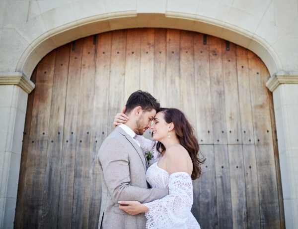 A couple portrait taken outdoors in front of a large arched wooden double door set within a stone archway. The bride and groom stand facing each other with foreheads touching, embracing closely. The bride wears an off-the-shoulder white lace dress with long sleeves and holds a small floral arrangement with soft pink blooms. The groom wears a light gray suit with a white shirt and no tie. The composition is a mid-length portrait shot with the arched doorway framing the couple symmetrically in the background.