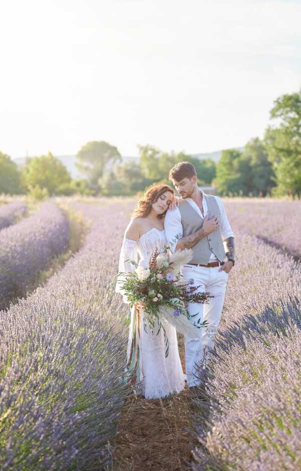 A couple portrait taken outdoors in a Provençal lavender field with rows of purple lavender extending behind them in warm golden-hour light. The bride wears an off-shoulder white lace boho-style dress with flowing sleeves and holds a large, loosely arranged bouquet featuring pampas grass, blush and ivory blooms, greenery, and trailing pastel ribbon streamers. The groom wears white trousers, a white shirt, and a gray vest with a brown leather belt, and has visible tattoos on his arms. The bride rests her head on the groom's shoulder as he looks down at her in a tender pose. The styling is distinctly boho with the relaxed lace gown, wildly arranged bouquet, and natural lavender field setting. Medium full-body portrait shot with soft backlit haze.