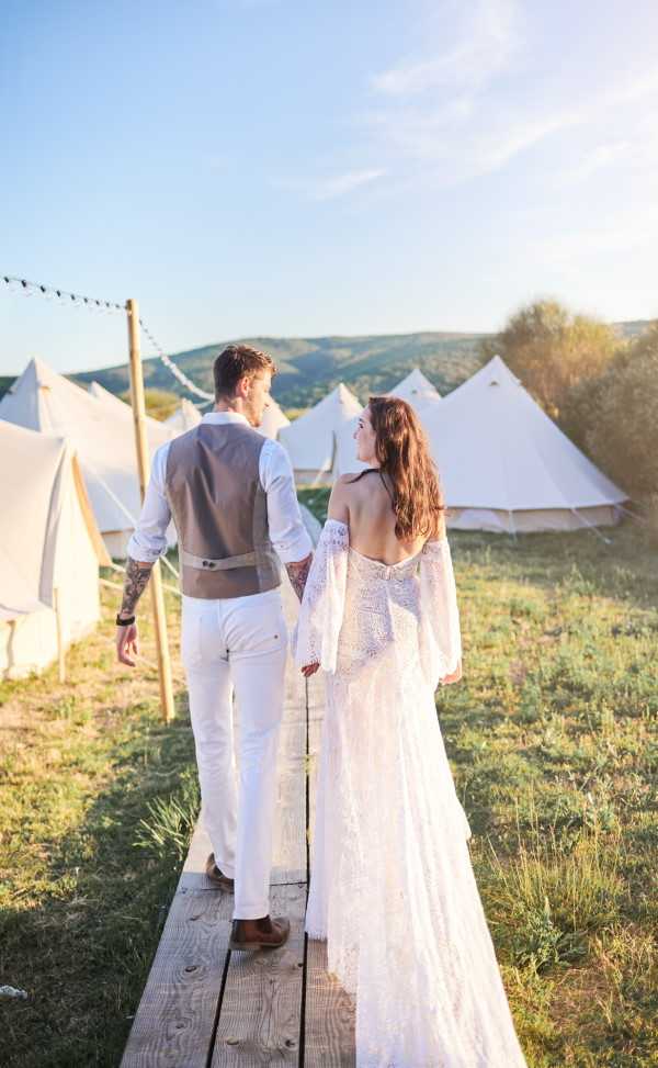 A couple walks hand-in-hand along a wooden boardwalk path through an outdoor glamping venue, photographed from behind at golden hour. The bride wears a backless white lace gown with long bell sleeves and a train, in a boho style, while the groom wears white trousers, a white shirt, and a tan waistcoat with visible tattoos on both arms. Multiple cream canvas bell tents are arranged across an open field in the background, with string lights strung between wooden posts. The composition is a full-length portrait shot with warm backlighting, giving a relaxed, festival-style boho wedding aesthetic.