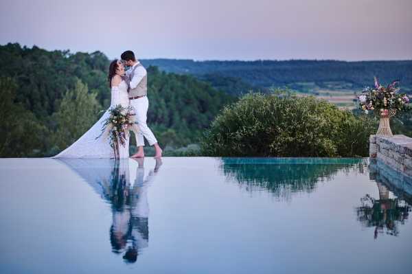 A couple portrait taken at dusk beside an infinity pool with a panoramic view over a wooded valley. The bride wears a long white lace dress and holds a large cascading bouquet featuring mixed wildflowers in warm tones of coral, burgundy, and greenery. The groom is dressed in a light grey suit with white shirt and no tie. The two stand barefoot at the pool's edge, embracing and about to kiss, with their full reflection mirrored in the still turquoise water. To the right, a rattan or gold-toned vase holds a tall floral arrangement in similar warm and muted tones. The styling leans toward a relaxed, bohemian-romantic aesthetic. Wide portrait shot with strong symmetry from the reflection. Potential venue feature image.