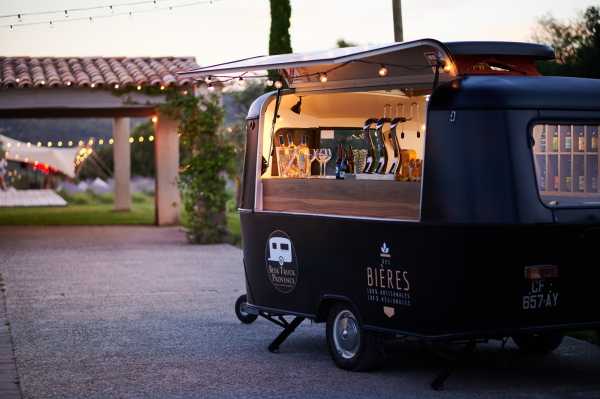 A mobile bar trailer, branded with 'Bières' and 'Mon Truck Provence' signage, is set up outdoors during what appears to be a cocktail hour at dusk. The matte black vintage-style trailer has a warm wood-paneled bar counter with the service hatch open, displaying wine bottles, champagne flutes, and glassware inside. String lights are strung in the background across a covered pergola area, adding warm ambient lighting to the early evening setting. The composition is a close-up three-quarter shot of the trailer, emphasizing it as a stylish, rustic-modern bar feature for the event.