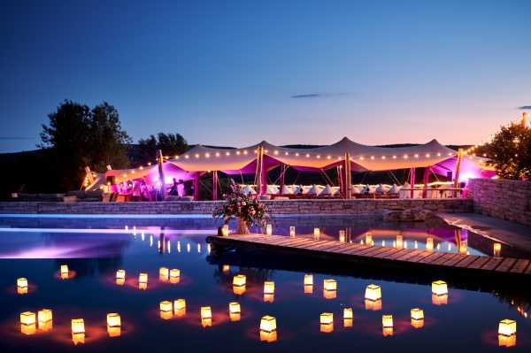 An outdoor evening wedding reception photographed in a wide shot at dusk, featuring a stretch tent structure with white draped canopy panels lit from within by warm pink and magenta uplighting, with festoon string lights looping along the tent's open sides. The reception tent appears to be set for a seated dinner with white-covered chairs and tables visible inside. In the foreground, a swimming pool reflects the warm golden glow of approximately 15-20 square floating lanterns arranged across the water's surface, creating symmetrical reflections. A wooden pool deck extends into the frame with a small floral arrangement and additional lanterns placed along it. The overall decor palette combines deep pink/magenta lighting with warm candlelight tones against the deep blue twilight sky. Potential venue feature image.