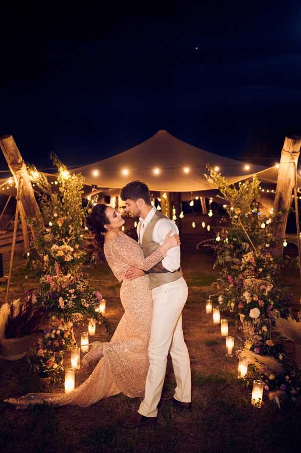 A couple embraces outdoors at night in front of a stretch tent reception marquee lit with festoon bulb lights. The bride wears a full-length long-sleeve gold sequined gown, and the groom wears cream trousers, a white shirt, and a taupe waistcoat. They are framed by two tall wooden post arrangements decorated with lush greenery and mixed florals in blush, mauve, and warm yellow tones. The ground around them is lined with glass pillar candles and low floral clusters in the same color palette. The warm candlelight and festoon lighting create a boho-rustic outdoor evening aesthetic. Wide portrait shot with the illuminated tent and dark night sky as the backdrop.