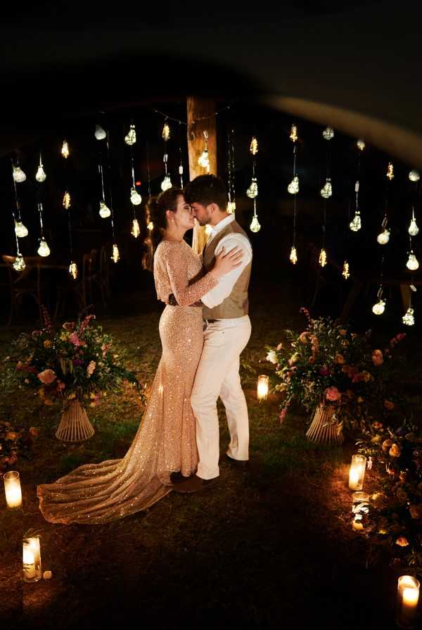 A couple shares an intimate moment outdoors at night, standing close together with their foreheads nearly touching in what appears to be a portrait or first dance setting. The bride wears a long-sleeve, form-fitting gold sequined gown with a trailing skirt, while the groom is dressed in cream trousers, a white shirt, and a tan vest. Behind them, a curtain of hanging Edison-style exposed bulbs is strung between wooden posts, creating a warm golden backdrop against the dark night sky. The ground around them is lined with glass candle votives and large floral arrangements in wicker or ribbed vases, featuring pink, magenta, and deep red blooms mixed with lush greenery, contributing to a romantic boho-rustic outdoor aesthetic. The composition is a medium portrait shot taken slightly from above, with the lighting almost entirely provided by the Edison bulbs and candles.
