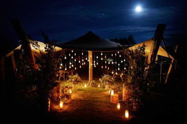 A wide-shot nighttime exterior image of an outdoor wedding ceremony space set beneath a large canvas or sailcloth tent structure with wooden timber frame posts. The entrance pathway is flanked by wooden crate planters filled with greenery and florals, with numerous candle lanterns placed along the ground on both sides creating a warm amber glow. A curtain of hanging pendant bulbs or globe lights is strung from the tent frame, creating a dramatic illuminated backdrop above the altar area. Additional floral arrangements are visible at the base of the posts. The overall decor palette is rustic-boho with warm candlelight tones against the deep blue night sky, which features a visible full moon. No people are present in the shot. Potential venue feature image.