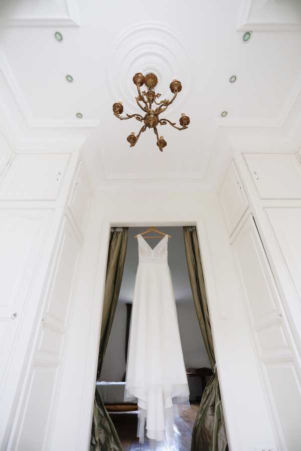 A getting-ready detail shot showing a white wedding dress hanging on a wooden hanger in a doorway between two rooms. The dress features a V-neckline, lace bodice, and a flowing chiffon skirt with a slight high-low or layered hem. The room has white paneled walls and ceiling with ornate molding, and a gold antique-style chandelier is prominently visible overhead. Olive green floor-length curtains frame the doorway on either side, and dark hardwood floors are visible in the background room. The composition is a wide, upward-angled shot that emphasizes the architectural details of what appears to be a classic French chateau or manor interior.