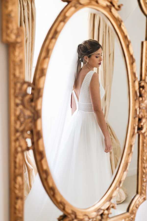 A getting-ready portrait of a bride reflected in a large ornate gold-framed oval mirror, shot indoors against a white wall with cream curtains visible in the background. The bride wears a white gown with a textured beaded or dotted bodice, a deep open back, and a full tulle skirt, paired with a long cathedral-length veil and drop earrings. Her dark hair is styled in a half-up updo, and she wears bold dark red lipstick. The composition is a medium portrait framed through the decorative gilded mirror, with a second mirror partially visible at the edge of the frame, creating a layered reflection effect.