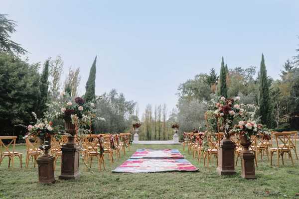 An outdoor ceremony setup photographed from the back of the aisle in a wide shot, showing a garden setting framed by tall cypress trees and mature greenery. The aisle is lined with natural wood cross-back chairs on both sides and marked by a colorful patchwork or kilim-style runner in pink, red, and blue tones. Four large ornate dark bronze or rust-colored pedestal urns anchor the entrance and mid-aisle points, each topped with lush floral arrangements featuring deep burgundy, blush pink, and cream blooms mixed with greenery and trailing foliage. At the far end of the aisle, two white stone pedestals hold additional floral arrangements, suggesting an altar area. The overall styling combines a boho and romantic aesthetic with rich jewel-toned florals, eclectic textile runner, and vintage-style urns against a natural garden backdrop.