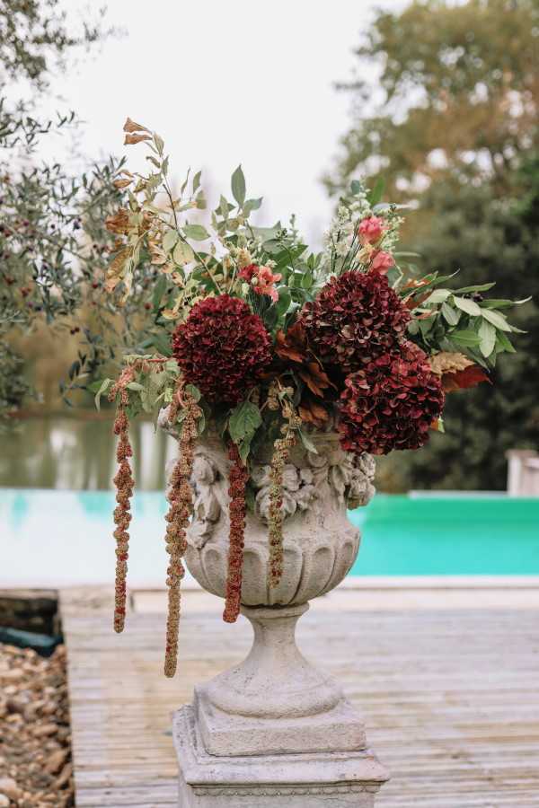 Close-up detail shot of a large ornate stone urn pedestal holding an oversized floral arrangement, positioned outdoors on a wooden deck beside a turquoise swimming pool. The arrangement features deep burgundy hydrangeas, blush pink blooms, trailing amaranthus in rust and green tones, and mixed greenery including eucalyptus and olive-like foliage, creating a rich autumn color palette. The carved stone urn with decorative relief detailing sits on a matching stone plinth. The overall floral styling is lush and organic with a moody, autumnal aesthetic.