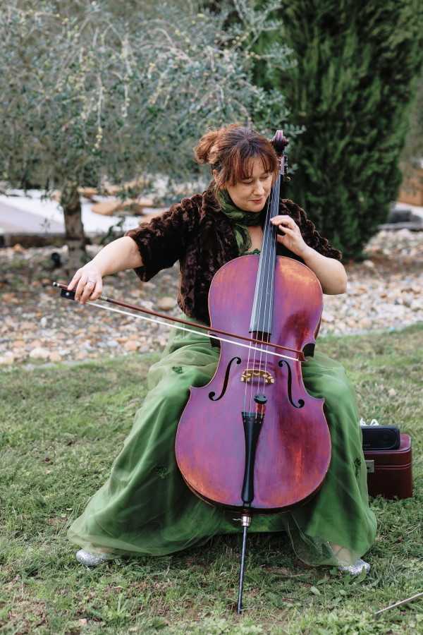 A female cellist performs outdoors at what appears to be a wedding cocktail hour or ceremony, seated on a chair in a garden setting with olive and cypress trees visible in the background. She is wearing a forest green full-length skirt, a dark brown faux-fur cropped jacket, and a green scarf, with silver glittery shoes visible beneath her skirt. She plays a warm reddish-brown cello, holding the bow mid-stroke, with a cello case visible on the ground to her right. The shot is a medium portrait framing the musician and her instrument in full.