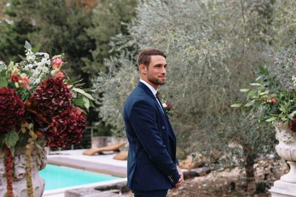 A groom stands outdoors during what appears to be a ceremony, positioned in profile and looking to the right of the frame. He is wearing a navy blue suit with a boutonniere featuring a deep burgundy flower. The setting is a garden or terrace area with a swimming pool visible in the background. Large stone urns flank the scene, filled with rich floral arrangements of deep burgundy hydrangeas, cream and white blooms, and greenery, consistent with an autumn color palette. The composition is a medium portrait shot with the groom as the focal point.