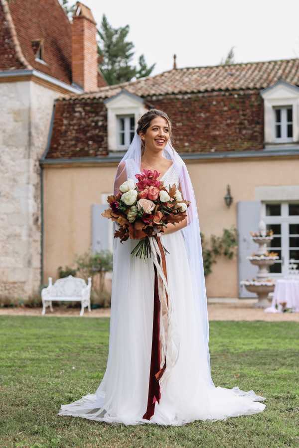 A full-length bridal portrait taken outdoors on the lawn of a French chateau with rendered stone walls, terracotta roof tiles, and grey-blue shutters. The bride wears a flowing white gown with a V-neckline, a long cathedral-length veil, and an updo hairstyle, and she is smiling broadly. She holds a large, loosely arranged bouquet featuring deep magenta orchids, ivory roses, blush roses, rust-toned dried leaves, and greenery, with long trailing ribbons in burgundy and blush. In the background, a white garden bench, a stone fountain, and a dessert display table with white linens are partially visible, suggesting a cocktail hour setup.