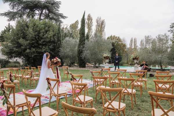An outdoor wedding ceremony is underway in a garden setting, with the bride walking down the aisle toward the groom who stands at the altar near a pool. The ceremony space is arranged with natural wood cross-back chairs in rows on either side of a colorful floral-printed aisle runner in pink, red, and green tones. The bride wears a white gown with a long veil and carries a large bouquet featuring deep red, burgundy, and warm-toned blooms. The groom stands in a navy suit at the far end of the aisle, accompanied by an officiant and a musician playing violin to the side. Floral arrangements with burgundy and warm-toned flowers also decorate the chair ends along the aisle. The overall styling is romantic and slightly bohemian, with a rich jewel-toned floral palette. This is a wide shot capturing the full ceremony layout from behind the bride.