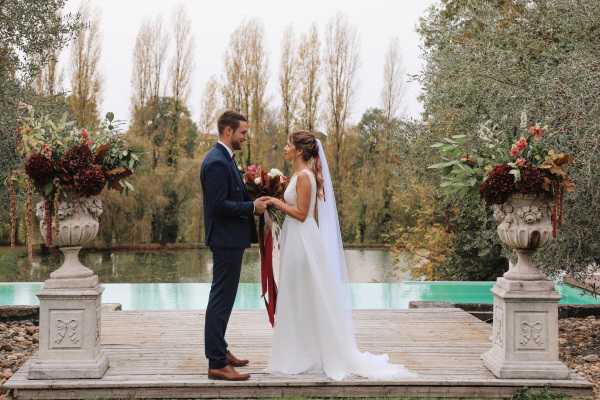 A bride and groom stand facing each other on a wooden deck platform in an outdoor setting beside a turquoise pool or lake. The groom wears a navy blue suit with brown leather shoes, while the bride wears a sleeveless, minimalist white gown with a long cathedral veil and holds a bouquet featuring deep burgundy flowers and red ribbon streamers. Two large stone urns on pedestals flank the couple, filled with rich autumnal arrangements of dark burgundy dahlias, coral blooms, and greenery. The composition is a full-length portrait shot with the couple centered, capturing an intimate moment as they hold hands and look at each other.