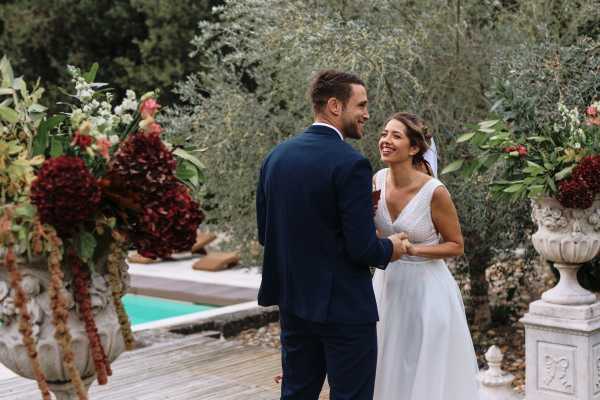 A bride and groom share a candid, laughing moment together during what appears to be an outdoor ceremony or portrait session, with a pool and wooden deck visible in the background. The groom wears a navy blue suit and the bride wears a white V-neck gown with cap sleeves and a short veil. Two large white stone urns flank the couple, filled with deep burgundy floral arrangements featuring dark foliage and rich red blooms. The shot is a medium portrait framed between the two decorated urns, with lush greenery in the background.