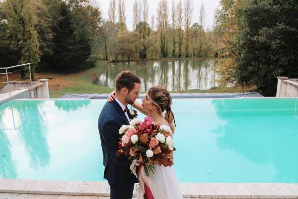 A couple portrait taken outdoors beside a turquoise swimming pool, with a calm lake and tall trees visible in the background. The groom wears a navy blue suit and the bride wears an off-the-shoulder white dress with her hair styled in an updo; they are facing each other in a near-kiss pose. The bride holds a large, lush bouquet featuring deep burgundy foliage, hot pink blooms, and ivory roses. The shot is a medium portrait composition with the pool and landscaped grounds serving as the backdrop.