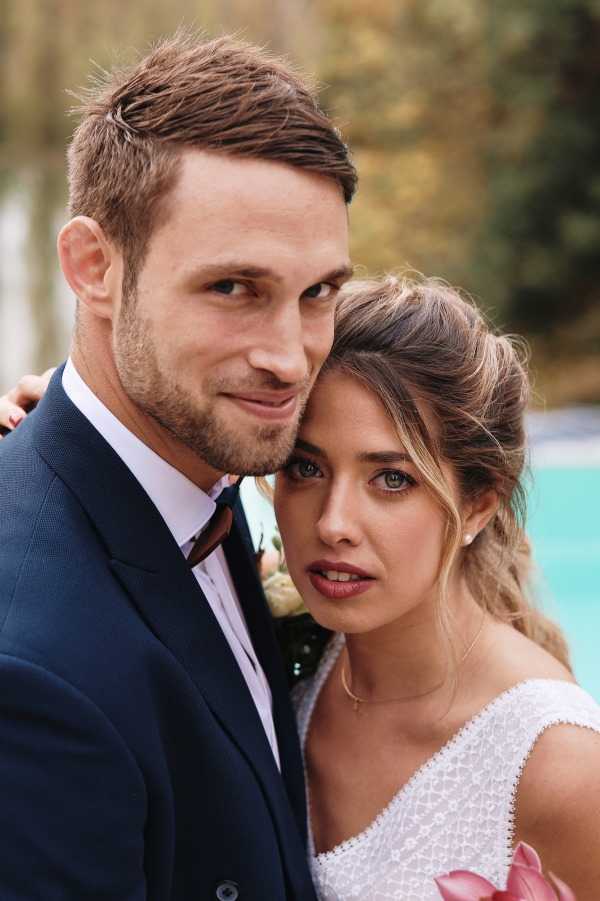 A close-up couple portrait taken outdoors, showing the bride leaning her head against the groom's cheek as both face the camera. The groom wears a navy blue suit with a white dress shirt and a burgundy tie, and has a small floral boutonniere visible on his lapel. The bride wears a white lace or textured sleeveless dress, has her dark brown hair styled in a loose updo, and holds what appears to be a pink floral bouquet partially visible at the bottom of the frame. A turquoise pool is softly blurred in the background, suggesting an outdoor venue with garden or estate grounds.
