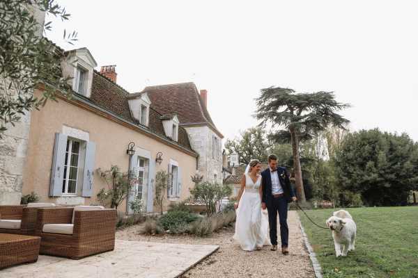 A bride and groom walk together along a gravel path beside a classic French manor house, accompanied by a large cream-colored dog on a leash. The bride wears a white lace V-neck gown and the groom is dressed in a navy suit with a bow tie. The manor features pale pink rendered walls, blue-grey shutters, and terracotta roof tiles, with rattan outdoor furniture visible on the terrace to the left. Wide shot captured from a slight distance, with a relaxed, candid feel during what appears to be a post-ceremony portrait session in the estate grounds. Potential venue feature image.