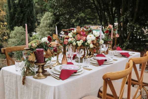 A styled outdoor reception tablescape set for approximately six guests, photographed in a wide angled close-up shot. The rectangular table is dressed with a white linen and features floral centerpieces combining ivory white peonies, deep burgundy blooms, blush pink tulips, and dried autumn leaves in brass and copper-toned vases. Place settings include stacked white charger plates with decorative pink-tinted inner plates, crystal glassware, and gold flatware, paired with folded burgundy napkins. Tall blush pink taper candles in brass candlesticks and a dark burgundy pillar candle add to the warm autumnal color palette, which mixes burgundy, blush, ivory, and gold tones in a romantic fall tablescape style. Natural wood cross-back chairs surround the table, set against a lush green garden backdrop.