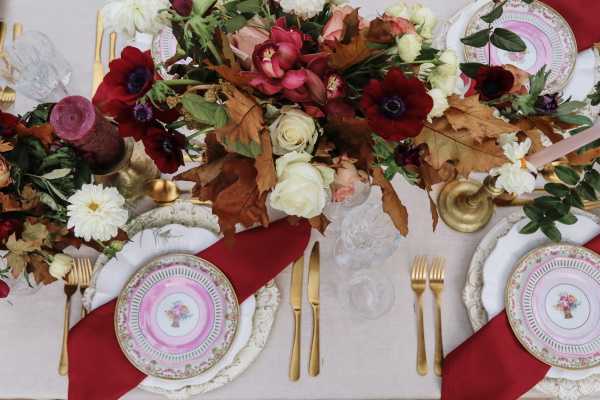 A close-up overhead shot of a wedding reception tablescape featuring vintage-style floral-rimmed plates with pink and gold detailing, layered on white lace charger plates. Gold flatware is paired with deep crimson linen napkins folded flat across each place setting. Crystal glassware is visible at the top of the settings. The centerpiece is a lush arrangement of deep burgundy and dark purple anemones, cream and blush roses, white dahlias, and dried autumn oak leaves in warm copper-brown tones, creating a rich fall color palette. Gold candlestick holders with dark wine-colored taper candles are placed along the table runner. The overall decor palette combines deep reds, burgundy, cream, and gold against a neutral linen tablecloth, with a classic-meets-autumnal styling theme.