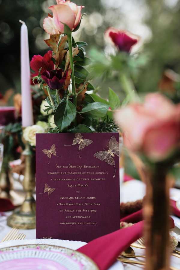 A close-up detail shot of a styled wedding tablescape featuring a deep burgundy wedding invitation with gold foil moth/butterfly illustrations, printed with formal wording for the marriage of Sally Megan to Michael Simon Jones at The Great Hall, Syon Park. The invitation is propped against a floral arrangement of burgundy anemones, blush and pink roses, cream florals, and green foliage. The table decor follows a rich jewel-toned palette with crimson napkins, gold cutlery, ornate gold candlestick holders with lavender taper candles, and decorative charger plates with gold and pink detailing. The overall styling theme is romantic and richly toned, combining deep berry, gold, and blush tones for a dramatic effect.