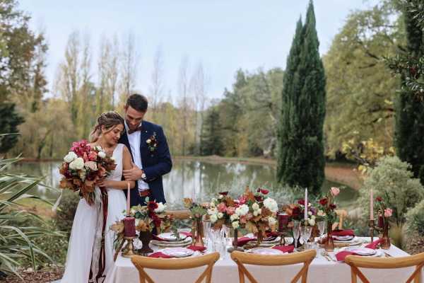 A couple poses together beside a styled outdoor reception table set in a garden with a pond visible in the background. The bride wears a sleeveless white gown and holds a large bouquet featuring deep burgundy, coral, blush, and ivory blooms mixed with dried autumnal leaves and long ribbon streamers; the groom wears a navy suit with a burgundy bow tie and boutonniere. The rectangular table is dressed with a white linen and set with natural wood cross-back chairs, burgundy napkins, gold-rimmed glassware, and layered place settings, with a lush floral runner of ivory garden roses, deep red dahlias, coral blooms, and rust-toned foliage running its full length, accented by tall dusty mauve taper candles in brass holders. The overall styling palette is autumn-romantic with deep burgundy, coral, ivory, and rust tones, and the image is a wide portrait-style shot capturing both the couple and the full table setting.