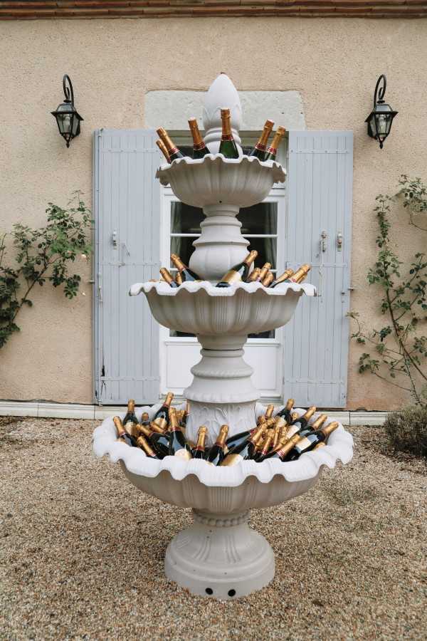 A cocktail hour or reception detail shot featuring a three-tiered white stone garden fountain repurposed as a champagne and wine bottle display. Each tier of the fountain is filled with multiple bottles bearing gold foil tops, arranged on ice. The fountain is positioned on a gravel surface in front of a pale render building facade with powder blue shutters and black iron wall lanterns on either side. The composition is a medium wide shot centered on the fountain. The styling is classic French countryside with a playful touch, using the architectural fountain as a functional drinks station.
