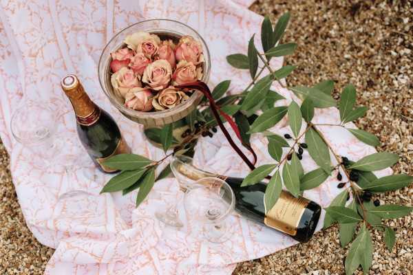 A styled flat lay detail shot arranged on a sandy outdoor surface, featuring a pink and white patterned fabric draped beneath the display. Two dark champagne bottles with gold and red foil labels are placed alongside two empty crystal champagne flutes. A glass bowl holds a cluster of blush and dusty rose garden roses tied with a deep burgundy ribbon, and an olive branch with dark berries extends across the right side of the composition. The overall palette is blush, gold, and deep green, suggesting a romantic, nature-inspired styling theme.