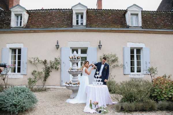 The bride and groom are performing a champagne tower pour outdoors in front of a French country manor house with pale stucco walls, blue-grey shutters, and a terracotta tile roof. The couple stands at a white linen-covered table holding a champagne tower of stacked glasses, with a decorative stone fountain visible just behind them. The bride wears a fitted white gown and the groom wears a navy suit with a bow tie. The composition is a wide portrait shot capturing both the couple and the full facade of the building. Potential venue feature image.