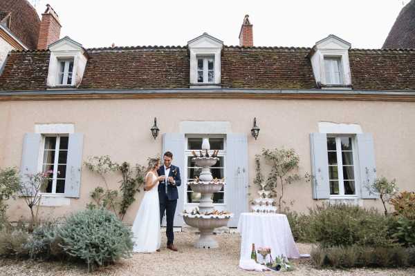 A bride and groom stand together on a gravel courtyard in front of a French country manor house with pale pink-beige rendered walls, blue-grey shutters, and terracotta roof tiles. The bride wears a white sleeveless fitted gown and the groom wears a navy suit with a white boutonniere; they appear to be pouring or examining champagne glasses. A champagne tower is displayed on a white linen-covered table to the right of the couple, with bottles of champagne visible at the base, and a large stone ornamental fountain sits directly behind them against the building facade. The shot is a wide portrait capturing both the couple and the full front elevation of the venue. Potential venue feature image.