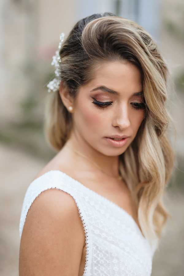 Close-up portrait of a bride looking downward, shot outdoors with a softly blurred background. She wears a white textured sleeveless dress with delicate beaded or lace detailing along the shoulder strap, and small pearl stud earrings. Her hair is styled in loose waves swept to one side, with small white floral hair pins — likely baby's breath or similar small blooms — tucked near the crown. Her makeup features a warm rosy-brown smoky eye with defined lashes and a neutral lip. The composition is a tight head-and-shoulders portrait with a shallow depth of field.