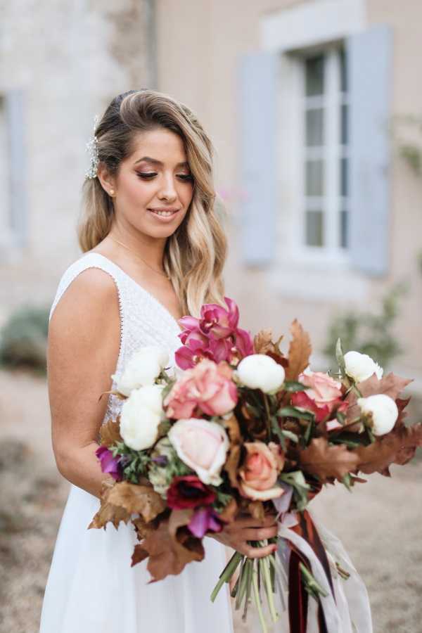 A bridal portrait taken outdoors in front of a French stone building with pale blue shutters, likely a chateau or manor house. The bride wears a sleeveless white V-neck dress with subtle texture and looks down at her bouquet, which is large and loosely arranged with blush and dusty pink roses, white ranunculus, deep magenta orchids, dark burgundy anemones, and dried autumn leaves in warm brown tones, tied with a dark ribbon in burgundy and forest green stripes. Her hair is worn half-up in loose waves with a delicate crystal or pearl hair accessory, and her makeup features warm, defined eye shadow and a natural lip. The composition is a close-up portrait framing the bride from roughly the waist up.