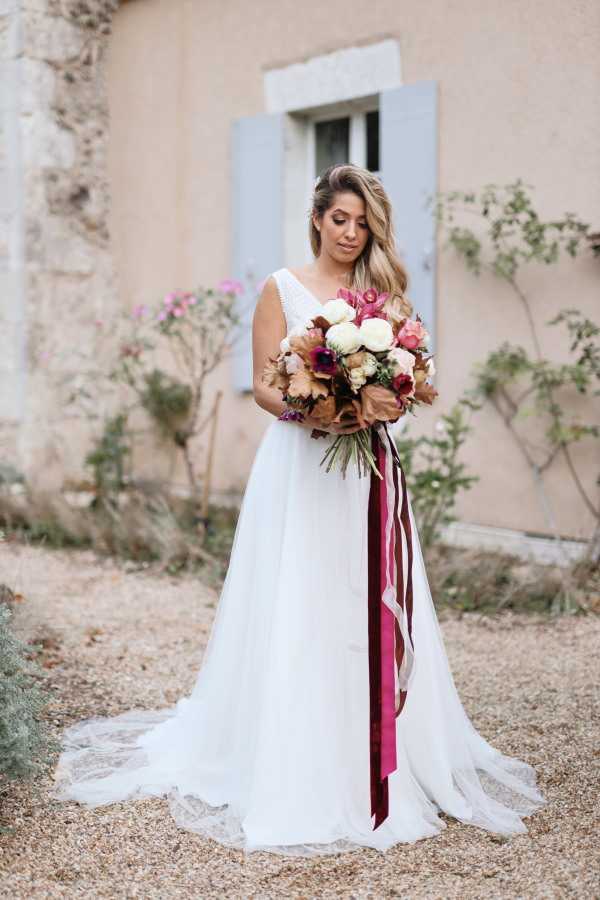 A bridal portrait taken outdoors on a gravel courtyard in front of a pale pink rendered building with a pale blue shutter window. The bride stands alone, looking down at her bouquet, wearing a white A-line gown with a V-neckline, thin straps with lace detailing, and a lace-edged train. Her blonde ombre hair is worn half-up with soft waves. She holds a rounded bouquet featuring ivory roses, burgundy anemones, dusty terracotta/copper ranunculus, and pink blooms, tied with long trailing ribbons in deep burgundy and blush pink. The overall styling leans romantic and loosely bohemian. Medium portrait shot with shallow depth of field, the background slightly blurred.