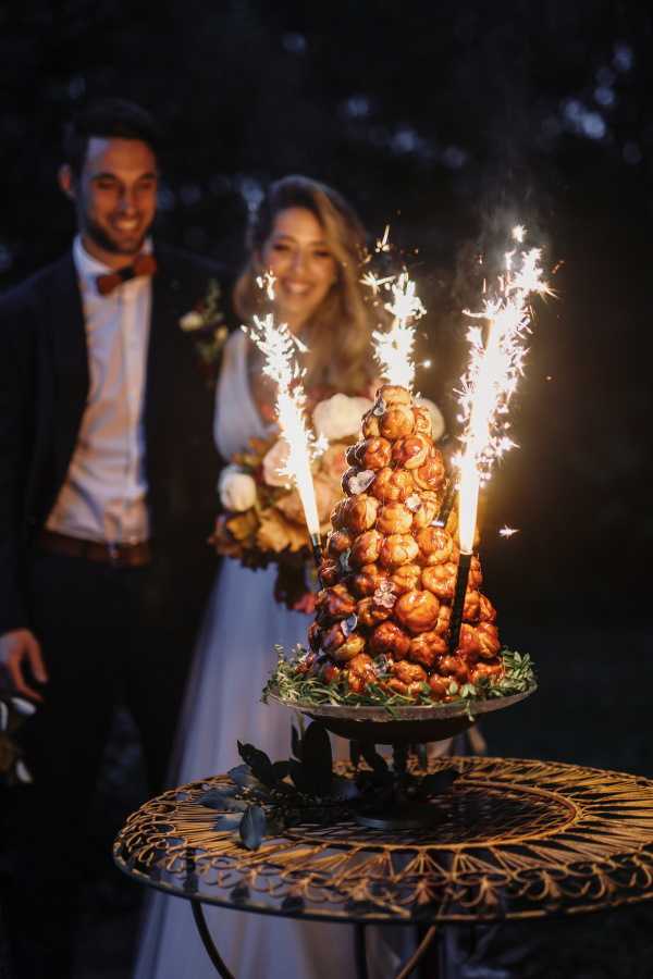 The couple stands behind a traditional French croquembouche during a nighttime cake presentation, with four lit sparklers shooting from the top of the choux pastry tower. The croquembouche is displayed on a decorative metal pedestal stand set on an ornate wrought-iron garden table, garnished with green herbs and dark foliage at its base. The groom wears a dark navy suit with a burnt orange bow tie, while the bride wears a white strapless gown and holds a bouquet featuring white blooms and warm autumn-toned florals. The portrait is a medium shot with the croquembouche in sharp focus in the foreground and the smiling couple intentionally soft-focused in the background, lit primarily by the warm glow of the sparklers against a dark outdoor setting.