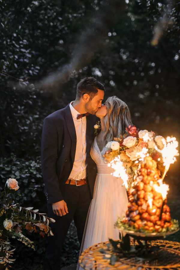 A couple shares a kiss outdoors at dusk or night in front of a croquembouche wedding cake adorned with ivory and blush roses, burnt orange flowers, and greenery, with sparklers actively firing from the base of the cake and casting warm golden light across the scene. The bride wears a white tulle skirt gown with a V-neck lace bodice and has loosely waved blonde hair, while the groom wears a dark navy suit with a burgundy bow tie, white shirt, and brown leather belt. A floral arrangement with blush roses and dark foliage is visible at the lower left. The composition is a medium portrait shot with the glowing cake and sparklers serving as the primary light source against a dark leafy background.