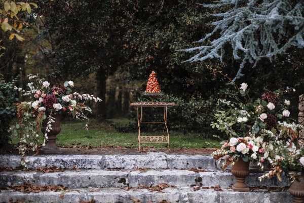 An outdoor garden cake display styled for a wedding, set on stone steps leading to a lawn surrounded by mature trees. A croquembouche — a traditional French tower of choux pastry puffs in warm caramel and red tones — sits atop a ornate vintage iron bistro table positioned at the base of the steps. Flanking the steps on either side are large terracotta urns overflowing with lush floral arrangements featuring blush and ivory roses, burgundy blooms, and trailing greenery and eucalyptus, creating an autumnal palette. The overall styling is romantic and classic French, with a moody, overcast atmospheric light and fallen leaves on the stone steps reinforcing the late-autumn seasonal feel. Wide establishing shot.