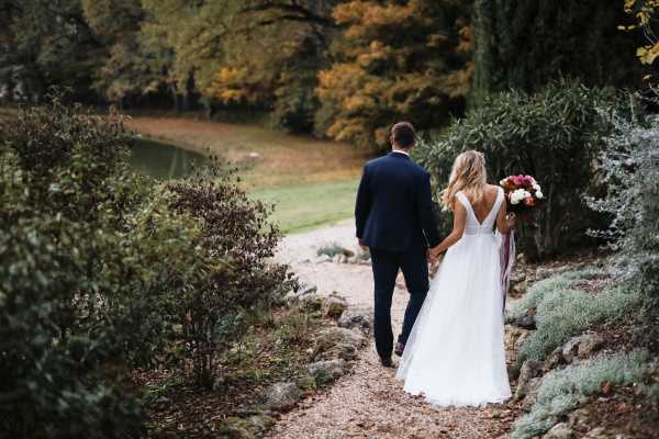 A couple portrait taken from behind as the bride and groom walk hand-in-hand along a gravel garden path in an outdoor setting with a small pond visible in the background. The groom wears a navy blue suit and the bride wears a white gown with a low V-back and a flowing skirt, carrying a bouquet of deep pink, coral, and white blooms with trailing burgundy ribbon. The autumn foliage in orange, yellow, and green tones frames the scene. The shot is a medium-wide portrait composition captured at ground level.