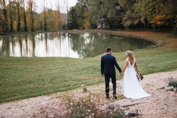 A bride and groom walk hand-in-hand toward a calm reflective pond on the grounds of what appears to be a French country estate, photographed from behind in a wide portrait shot. The groom wears a navy suit and the bride wears a white gown with an open low back and a flowing A-line skirt, carrying a small bouquet with deep burgundy and purple tones. The setting is outdoor with an autumn atmosphere, indicated by golden and orange foliage on the surrounding trees. The composition is a full-length rear-facing shot emphasizing the couple's movement through the landscaped grounds.