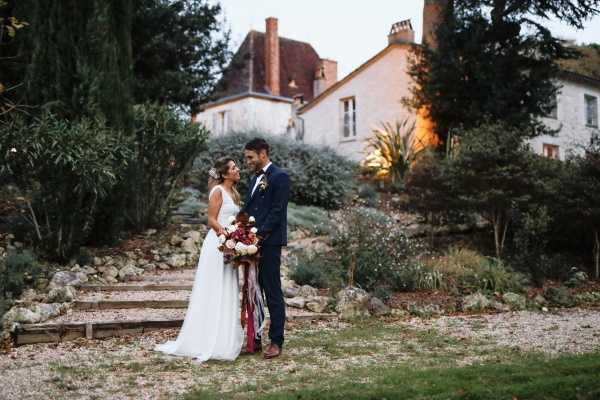 A couple portrait taken at dusk in the landscaped garden of a French chateau or manor house, with the lit building visible in the background. The bride wears a simple white sleeveless gown with a short train, her hair up, and holds a large bouquet featuring deep burgundy and blush blooms with long trailing burgundy and red ribbons. The groom wears a navy suit with a bow tie and burgundy shoes. The two stand close together, facing one another, on a gravel path at the base of stone garden steps. The overall styling is romantic and autumnal with a rich jewel-toned color palette. Medium-distance portrait shot.