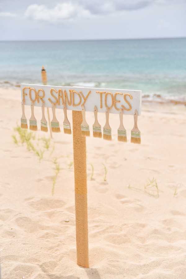 A close-up detail shot of a beach wedding decor element: a white wooden sign reading 'FOR SANDY TOES' in gold rope lettering, mounted on a rope-wrapped stake planted in the sand. Approximately ten small paintbrushes are hung along the bottom edge of the sign, intended for guests to brush sand off their feet. The sign is positioned at the beach ceremony or guest arrival area, with the ocean visible in the soft-focus background. The styling is casual coastal, with natural rope and neutral tones consistent with a relaxed beach wedding aesthetic.
