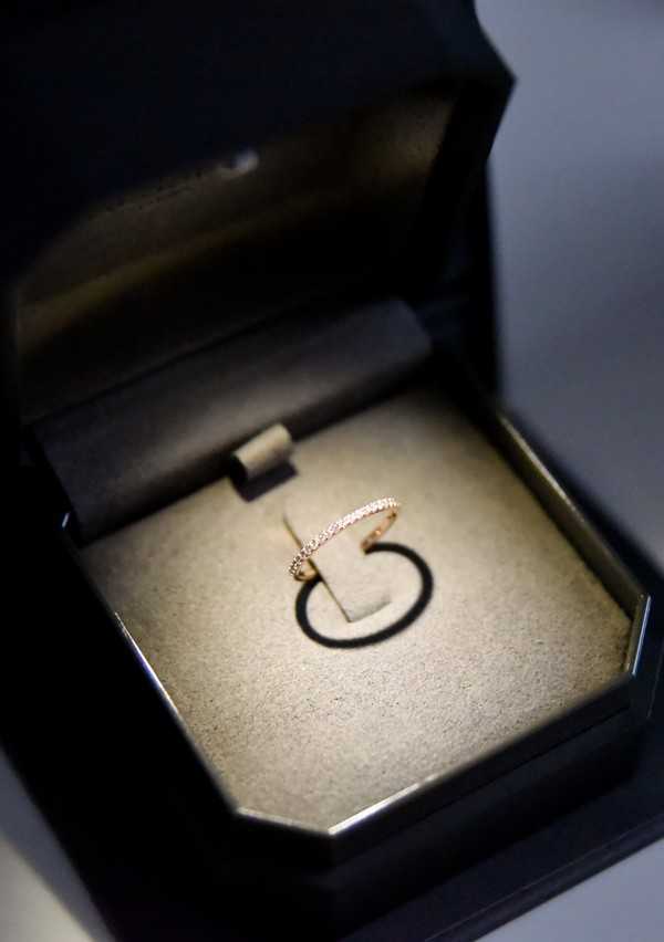 Close-up detail shot of a rose gold eternity band set with small pavé diamonds, displayed upright in an open black jewelry box with a cream velvet interior. The ring is slim and delicate, fully encircled with stones, and is spotlit against the pale cushioned lining, casting a circular shadow. The box has a dark lacquered octagonal exterior with a black satin lid lining. No people are visible; this is a ring detail image likely taken during the getting-ready or flat-lay portion of the wedding day.