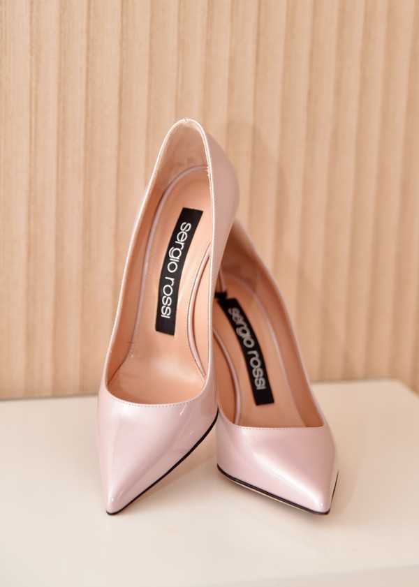 Close-up detail shot of a pair of Sergio Rossi blush pink patent leather pointed-toe stiletto heels, placed upright and leaning against each other on a white surface. The shoes have a high stiletto heel with a metallic tip and black-branded insoles clearly displaying the Sergio Rossi logo. The getting-ready detail style is clean and minimal, with the shoes positioned against a light wood-paneled background.