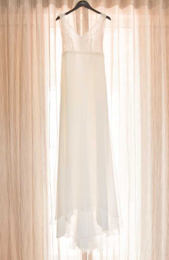 A getting-ready detail shot of a white wedding dress hanging on a black hanger against sheer, warm-toned curtains backlit by natural light. The gown features a V-neckline bodice with sequin or lace embellishment on the upper portion, transitioning to a smooth, flowing chiffon or crepe skirt with a subtle layered hem. The dress has a sleeveless, minimalist silhouette with a slightly asymmetric handkerchief-style hemline. The composition is a full-length vertical shot centered on the dress.