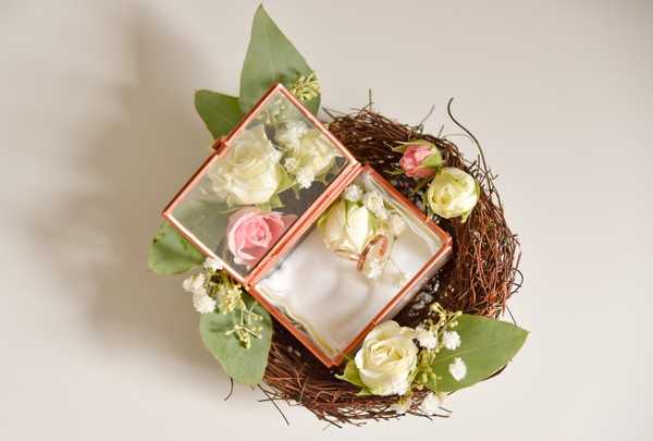 Close-up flat lay detail shot of a rose gold geometric glass ring box displayed open on a rustic twig nest, shot against a plain light grey background. Two gold wedding bands rest on a white cushion inside the box, which is decorated with cream and pink spray roses, baby's breath, and green leaves arranged around and inside the lid. The styling is romantic and garden-inspired, combining the organic texture of the woven twig nest with the clean lines of the copper-framed glass box.